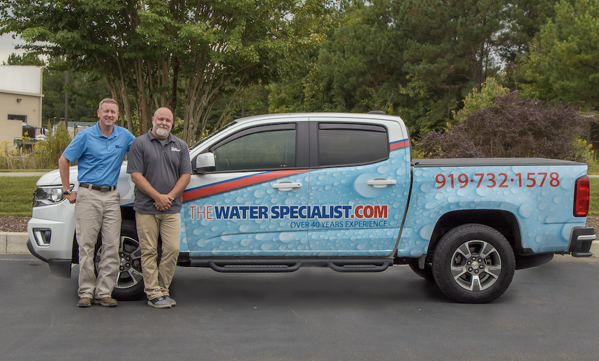 Two men standing in front of a pickup truck with The Water Specialist logo and artwork